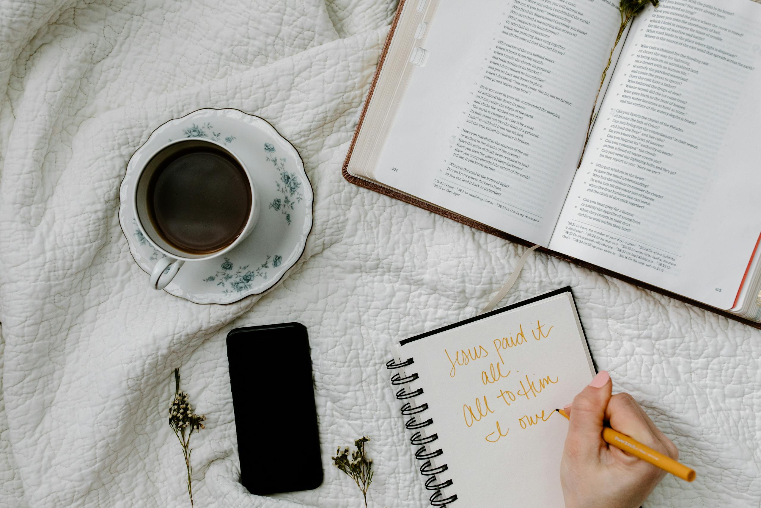 Flat lay of Bible study with coffee, notebook, and phone on a quilted blanket.